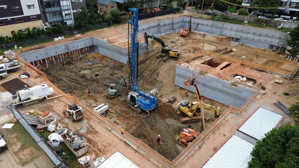 Aerial view of a basement excavation site featuring concrete retaining walls, a Soilmec SR-45 piling rig, excavators, and foundation plant machinery.