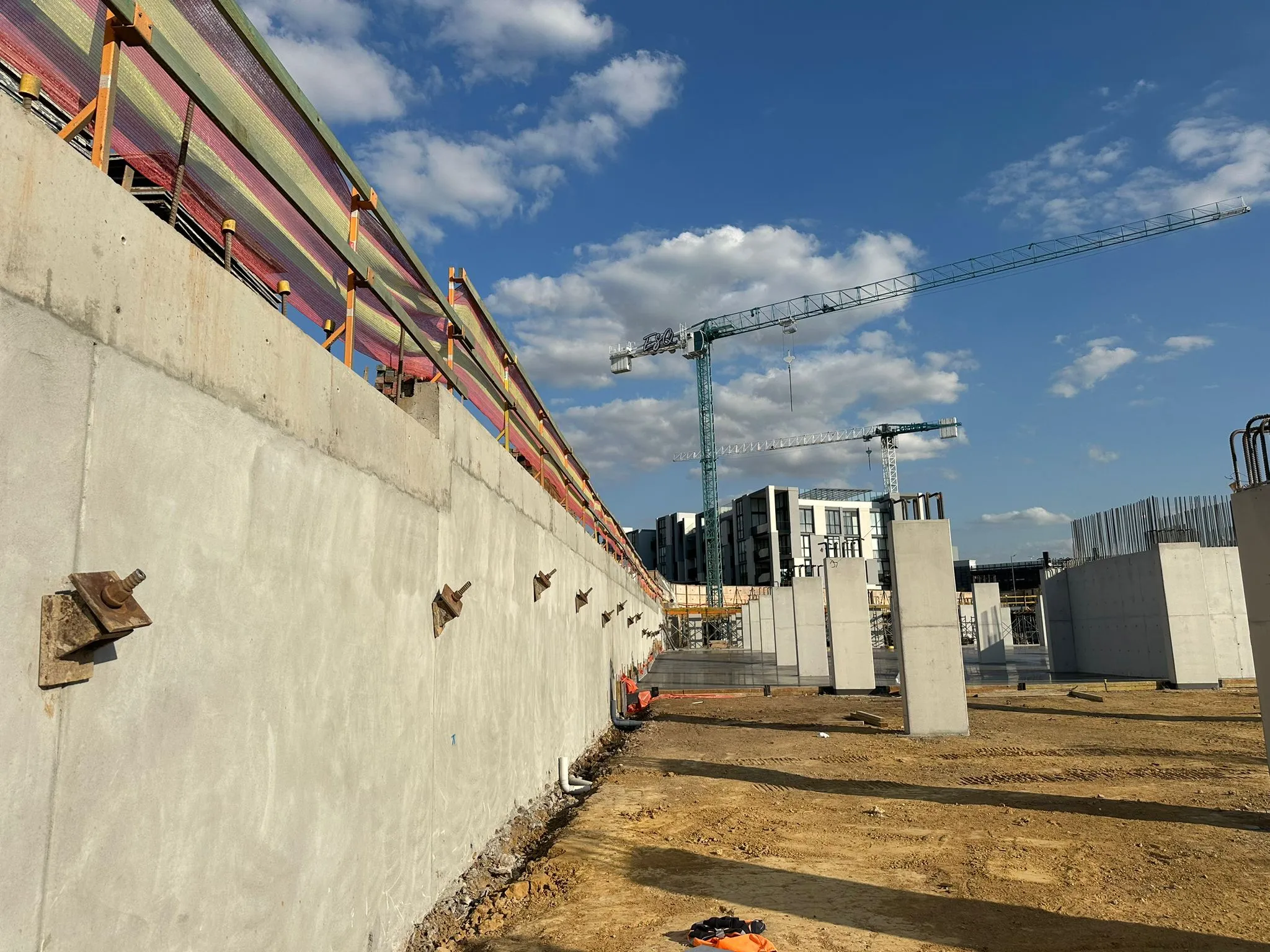 Concrete retaining wall fitted with post-tensioned ground anchor heads alongside concrete columns and tower cranes on a construction site.