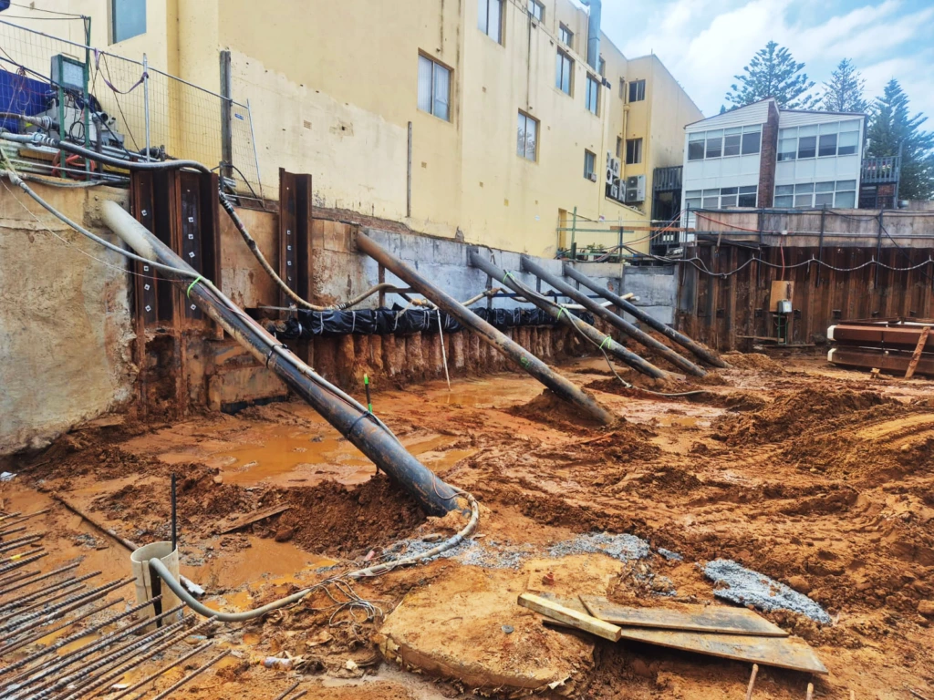 Heavy-duty diagonal steel props supporting a retaining wall during deep basement excavation next to existing multi-storey buildings.