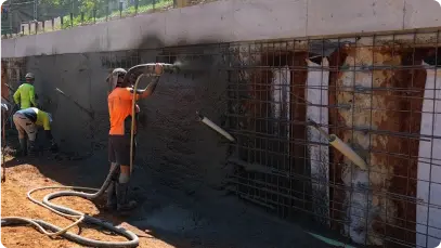 A nozzleman spraying wet-mix shotcrete over steel reinforcement mesh onto a basement retaining wall face while crew members finish the concrete surface.