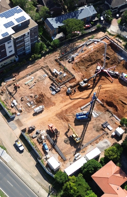 Aerial perspective of a large civil engineering site surrounded by residential buildings, featuring heavy machinery, excavators, and concrete supply trucks.