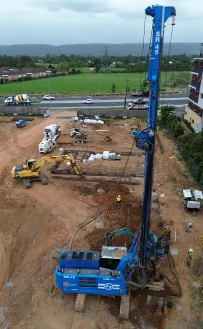 Aerial drone shot of a Soilmec SR-45 piling rig with mast extended, surrounded by excavators and concrete mixer trucks on a construction site.