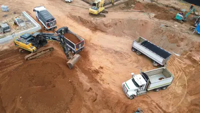 Heavy tracked excavator loading soil alongside truck and dog haulage trailers on a civil site.