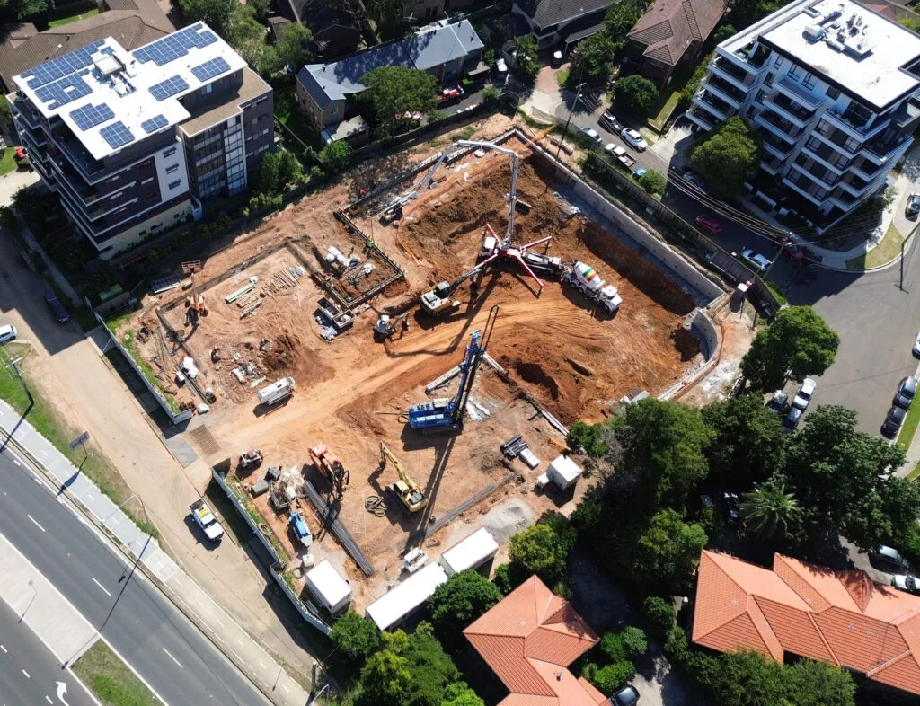 Aerial drone photograph of an urban construction site showing bulk excavation, earthmoving equipment, a concrete pump boom, and foundation piling