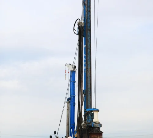A blue Soilmec SR-45 piling rig with a tall vertical mast and a Bobcat loader operating on an open construction site.