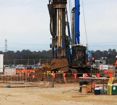 A blue Millent drilling rig discharging soil from a large auger attachment on an open construction site.
