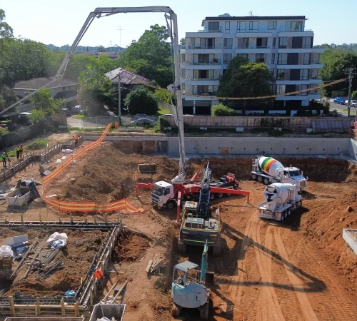 High-angle drone photo of a mobile concrete boom pump and agitator trucks conducting a slab foundation pour on an excavated construction site