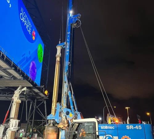 A blue Soilmec SR-45 piling rig with a raised vertical mast operating next to a large illuminated digital billboard at night.