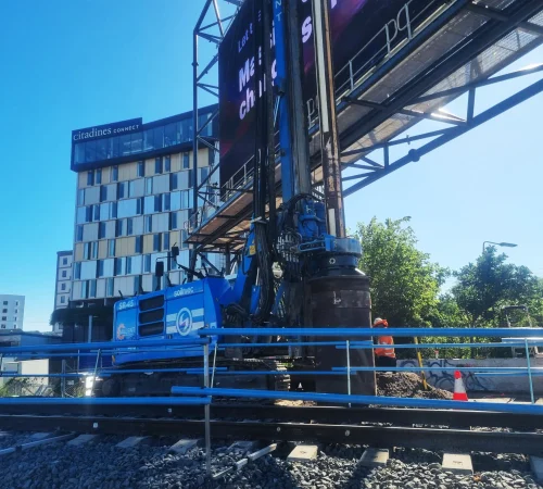 A blue Soilmec SR-45 piling rig operating next to railway tracks with the Citadines Connect hotel in the background.