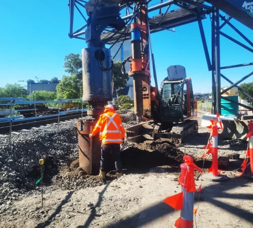 Construction worker in hi-vis standing next to an excavator-mounted drill attachment near railway tracks.