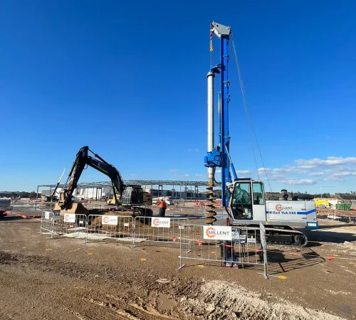 A Soil Tek S60 drilling rig with an exposed auger standing alongside an excavator on a clear, open construction site.