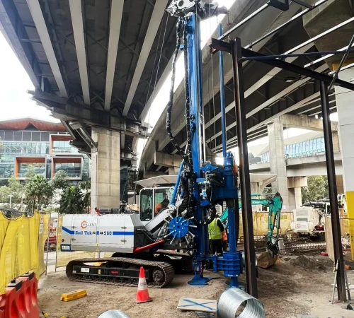 Compact Soil Tek S60 piling rig operating under an elevated overpass bridge on a restricted urban construction site.