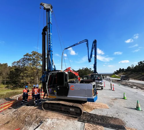 Millent foundation piling rig and concrete boom pump operating alongside construction workers on a road infrastructure project.