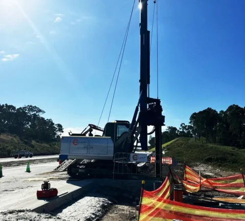 Track-mounted drilling rig set up next to temporary safety fencing at a civil roadwork construction site.