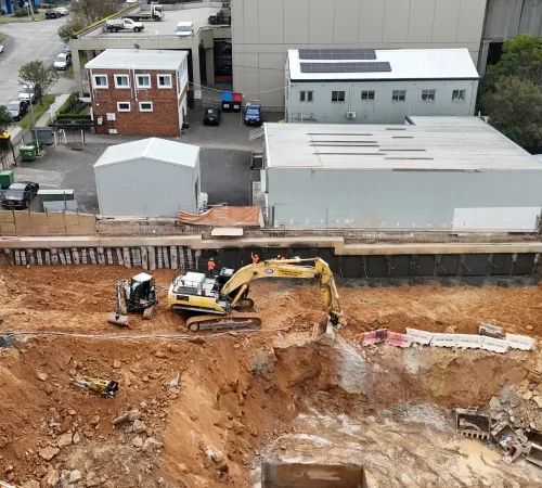 Overhead view of a bored pile shoring wall with shotcrete panels, a capping beam and two excavators working in the cut below
