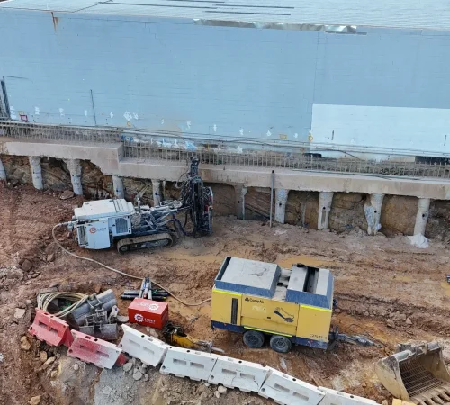 Tracked anchor drilling rig working below a capping beam and bored piles, with rock exposed between them and a building above