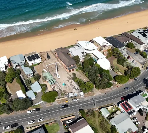 Aerial view of a piling rig and excavators on a narrow beachfront block between neighbouring homes, beside the beach and coastal road