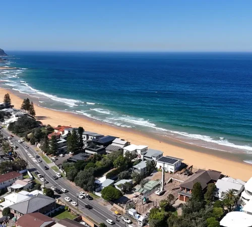 Aerial view along a Central Coast beach with the piling site among beachfront homes and the coastal road running behind