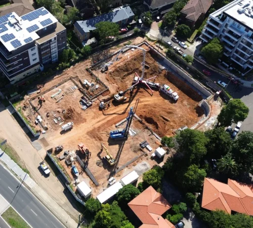 Aerial drone photograph of an urban construction site showing bulk excavation, earthmoving equipment, a concrete pump boom, and foundation piling